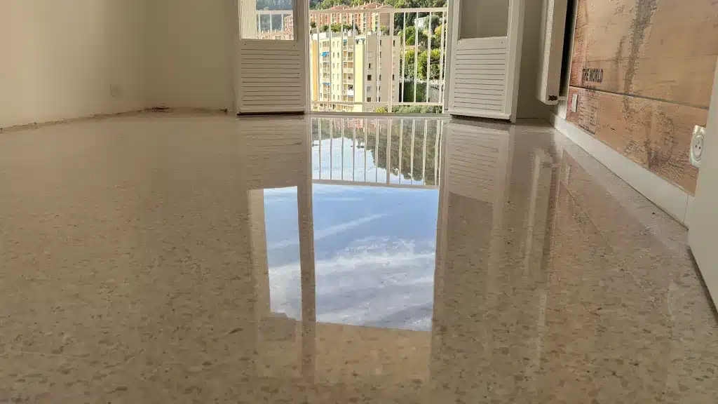 poncage-renovation-granito-terrazzo-grasse Chambre d'un appartement de Grasse avec un sol en granito poli-brillant reflétant la porte-fenêtre, le balcon et le ciel, avec vue sur les immeubles voisins