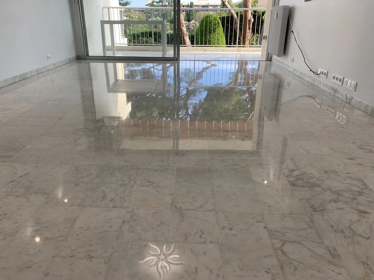 marbre-gris-clair-veine-poli-brillant-sejour-reflet-baie-golfe-juan Séjour d'un appartement de Golfe-Juan avec un sol en marbre veiné gris clair poli-brillant, reflétant la baie vitrée, le balcon, les pins et un luminaire du plafond